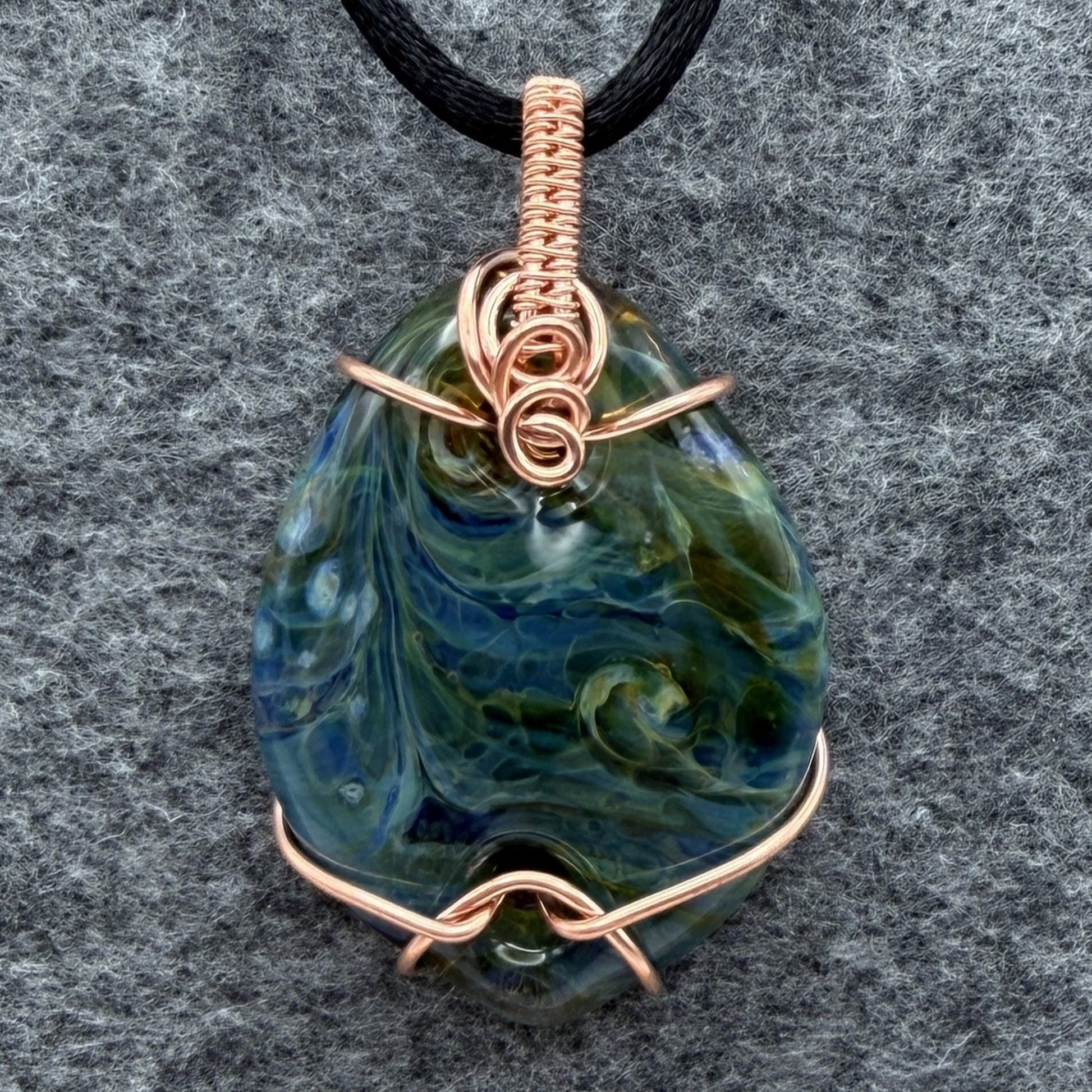 Front view of side 2 of a handmade pendant necklace featuring a marbled blue and green glass stone wrapped in copper wire, hanging from a black cord against a textured gray background.