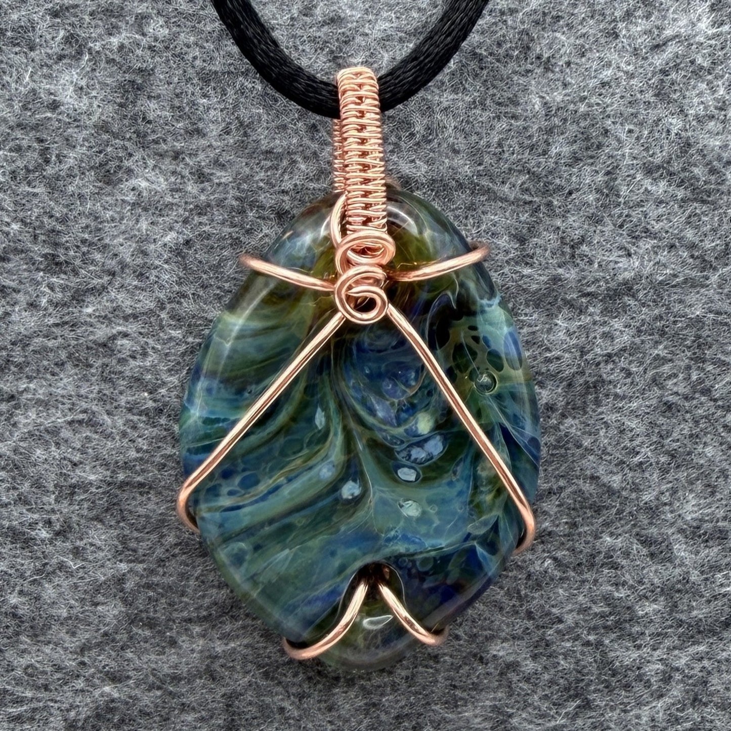 Front view of side 1 of a handmade pendant necklace featuring a marbled blue and green glass stone wrapped in copper wire, hanging from a black cord against a textured gray background.