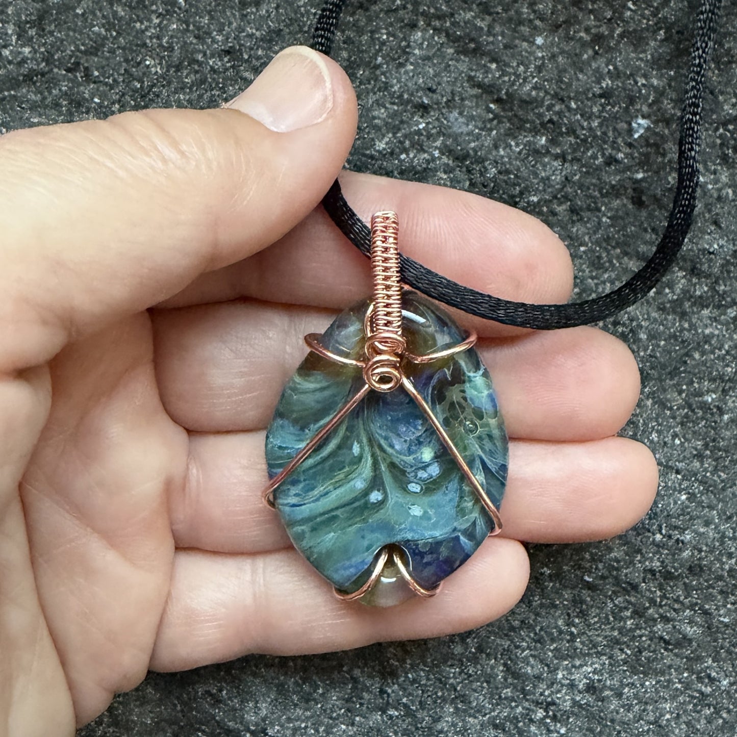 Front view of side 1 of a handmade pendant necklace featuring a marbled blue and green glass stone wrapped in copper wire, hanging from a black cord held in the artist's hand.