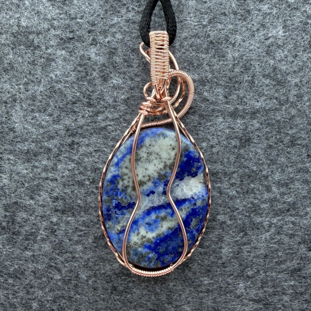 Back view of a teardrop-shaped lapis lazuli pendant with a copper wire wrap and a black cord, displayed on a textured gray background.
