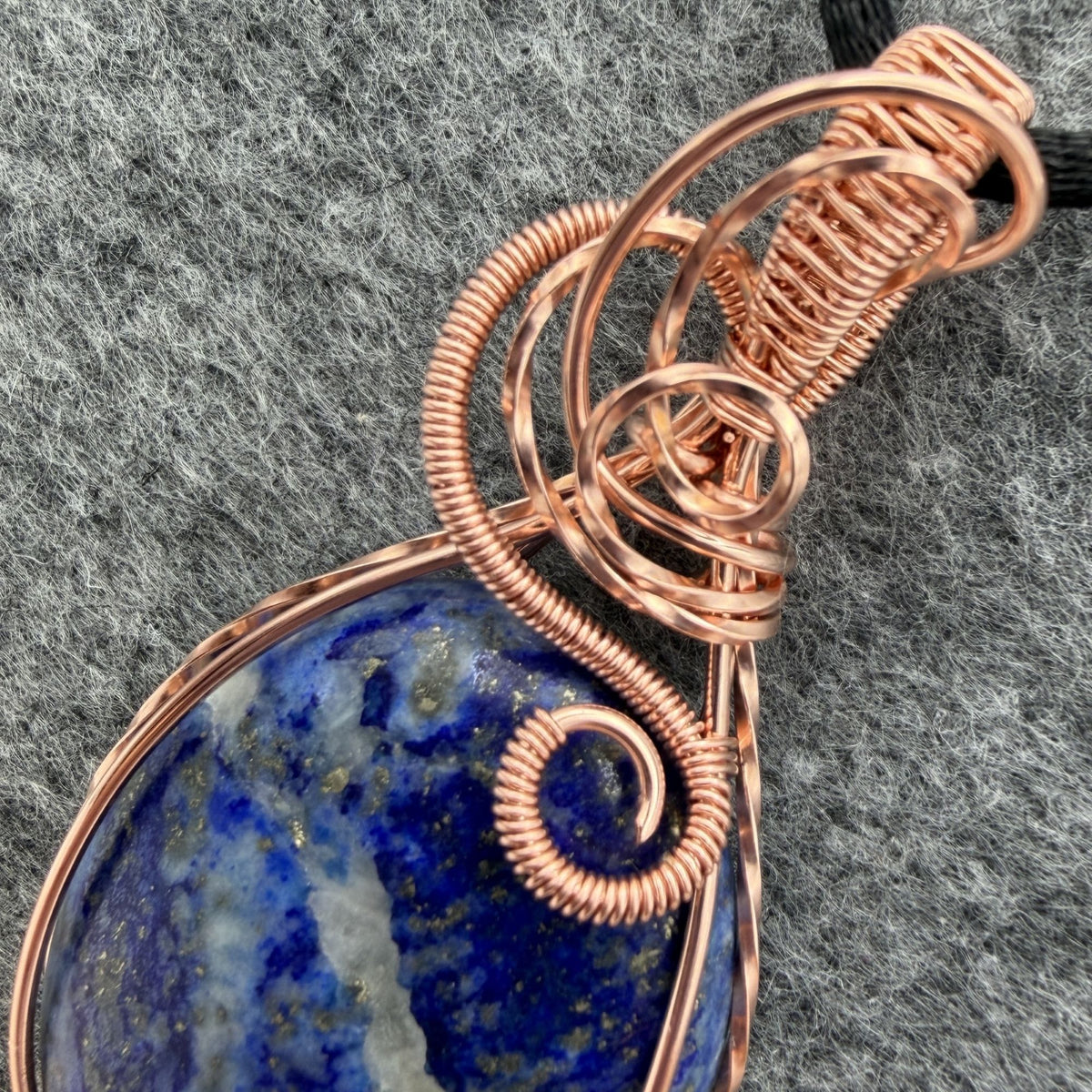 Detail view of a teardrop-shaped lapis lazuli pendant with a copper wire wrap and a black cord, displayed on a textured gray background.