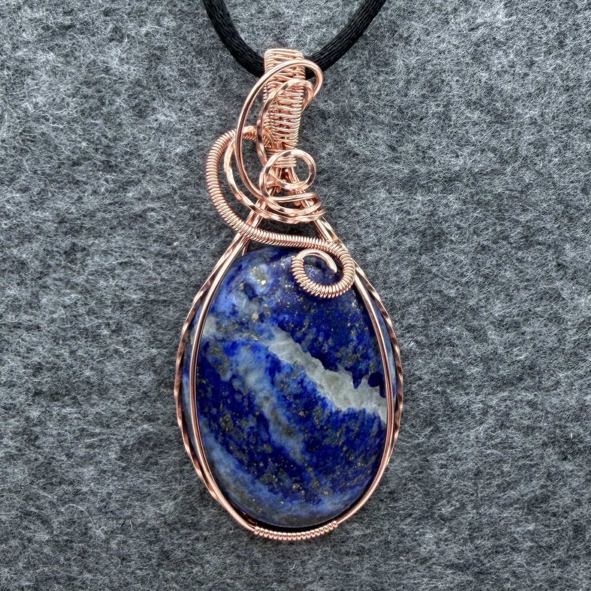 Front view of a teardrop-shaped lapis lazuli pendant with a copper wire wrap and a black cord, displayed on a textured gray background.