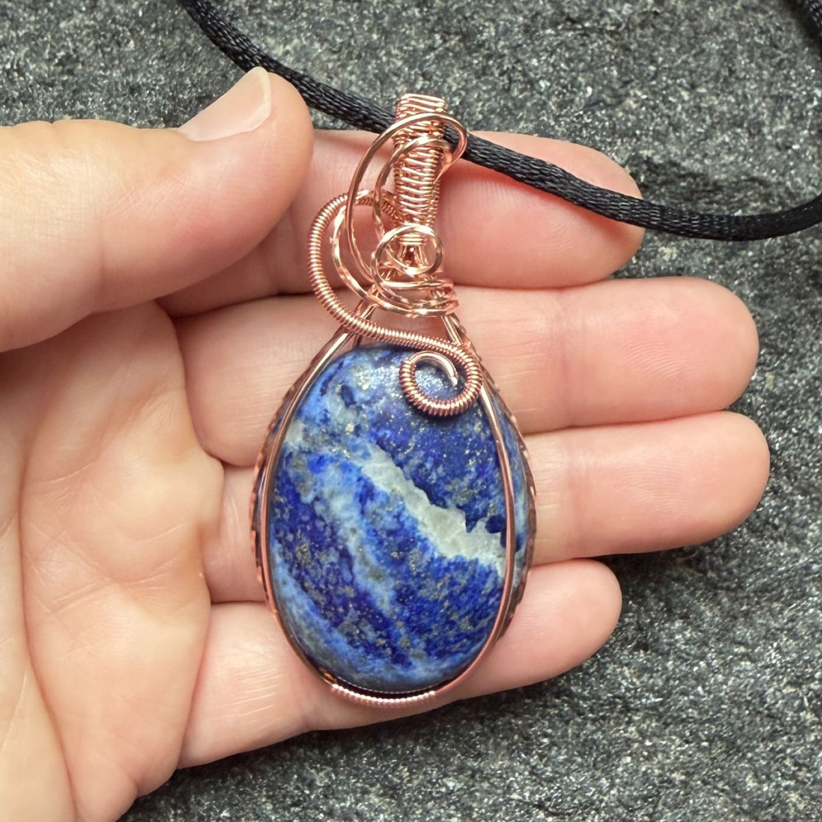 Front view of a teardrop-shaped lapis lazuli pendant with a copper wire wrap and a black cord, held in the artist's hand.