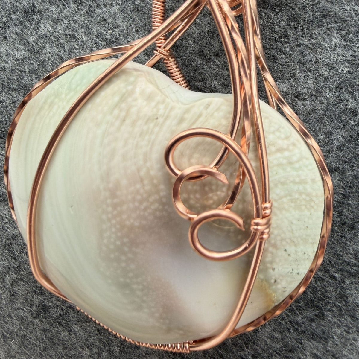 Detail view of side two of a handmade, heart-shaped pendant necklace featuring a sage and pink tinted seashell, wrapped in copper wire, hanging from a black cord against a textured gray background.