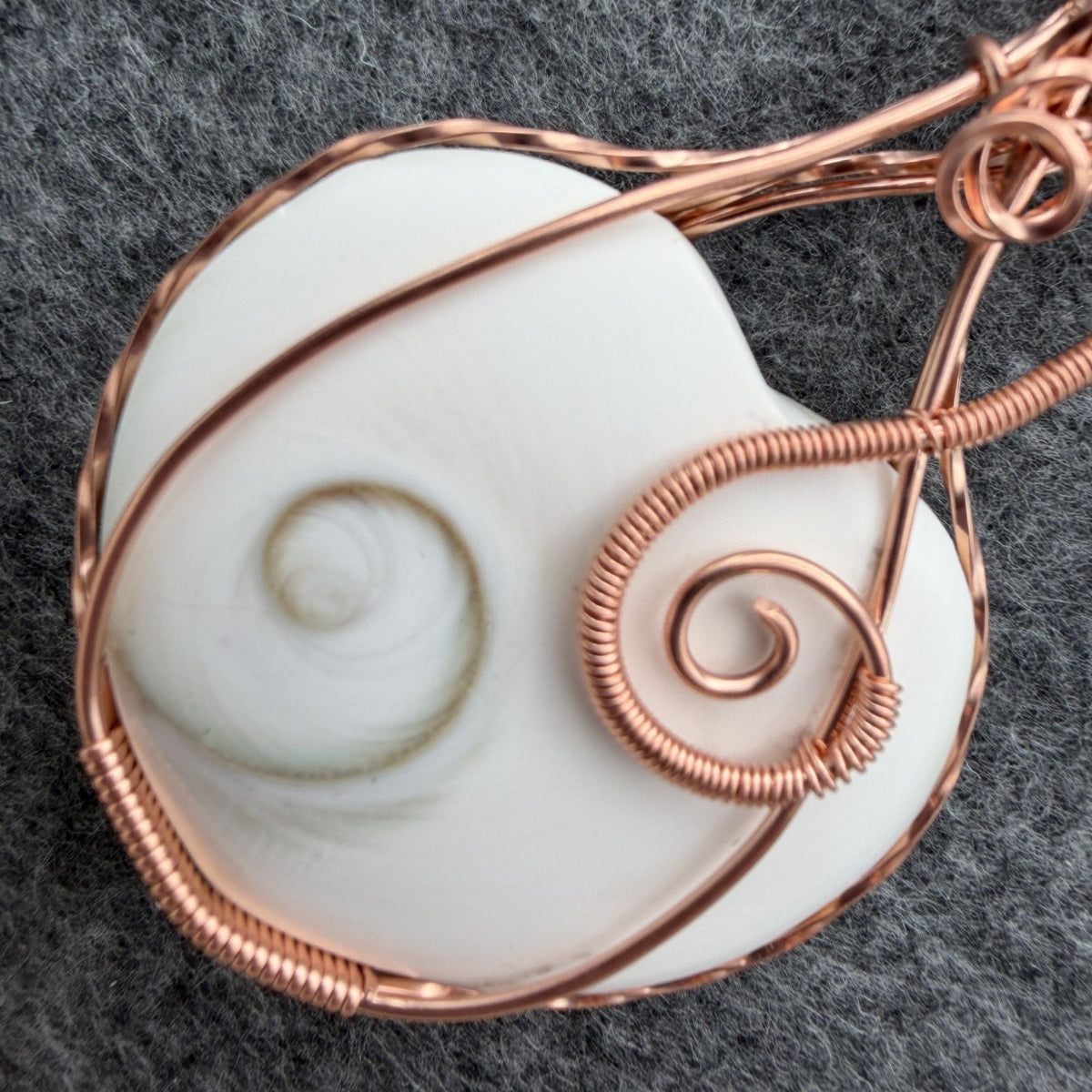Detail view of side one of a handmade, heart-shaped pendant necklace featuring a white seashell with a spiral pattern, wrapped in copper wire, hanging from a black cord against a textured gray background.
