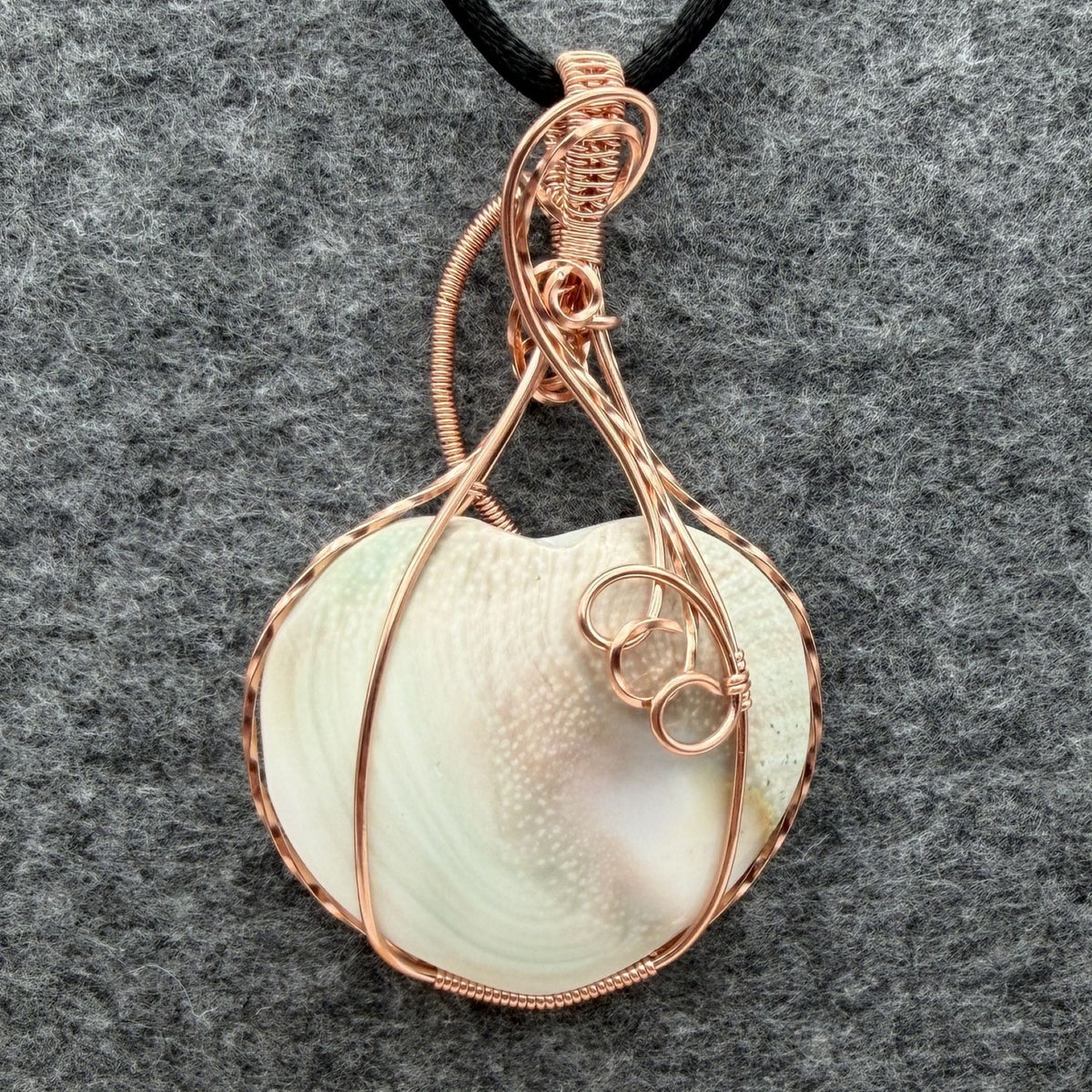 Front view of side two of a handmade, heart-shaped pendant necklace featuring a sage and pink tinted seashell, wrapped in copper wire, hanging from a black cord against a textured gray background.