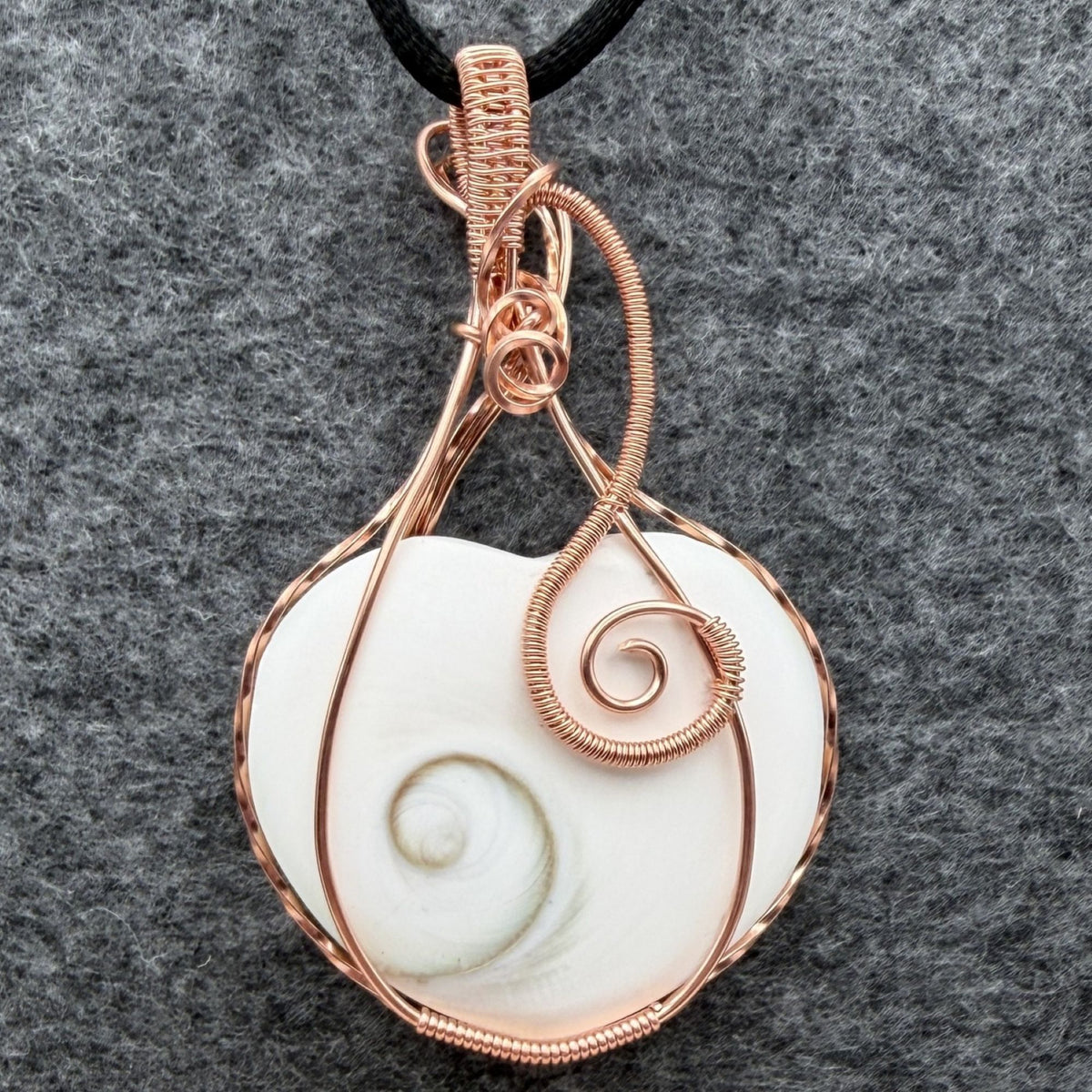Front view of side one of a handmade, heart-shaped pendant necklace featuring a white seashell with a spiral pattern, wrapped in copper wire, hanging from a black cord against a textured gray background.