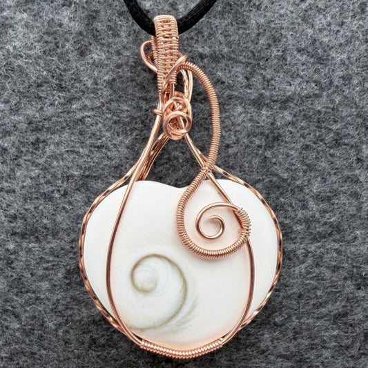 Front view of side one of a handmade, heart-shaped pendant necklace featuring a white seashell with a spiral pattern, wrapped in copper wire, hanging from a black cord against a textured gray background.