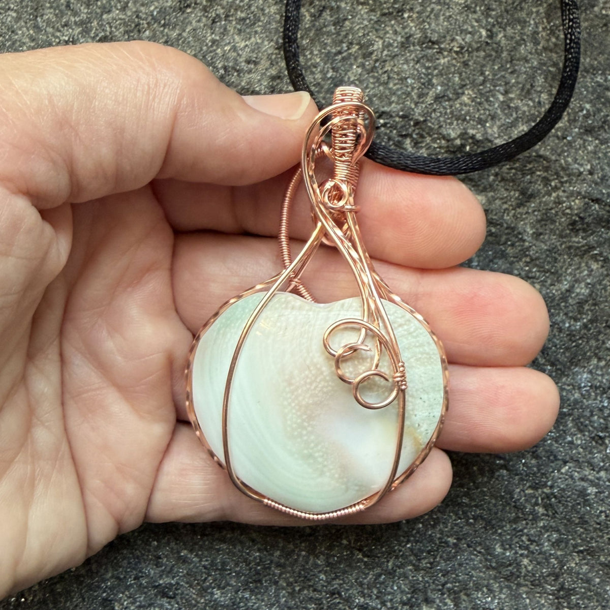 Front view of side two of a handmade, heart-shaped pendant necklace featuring a sage and pink tinted seashell, wrapped in copper wire, hanging from a black cord and held in the artist's hand.