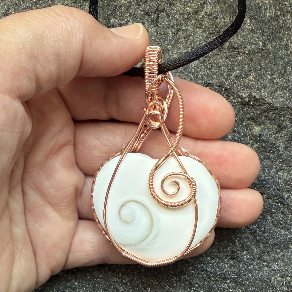 Front view of side one of a handmade, heart-shaped pendant necklace featuring a white seashell with a spiral pattern, wrapped in copper wire, hanging from a black cord and held in the artist's hand.