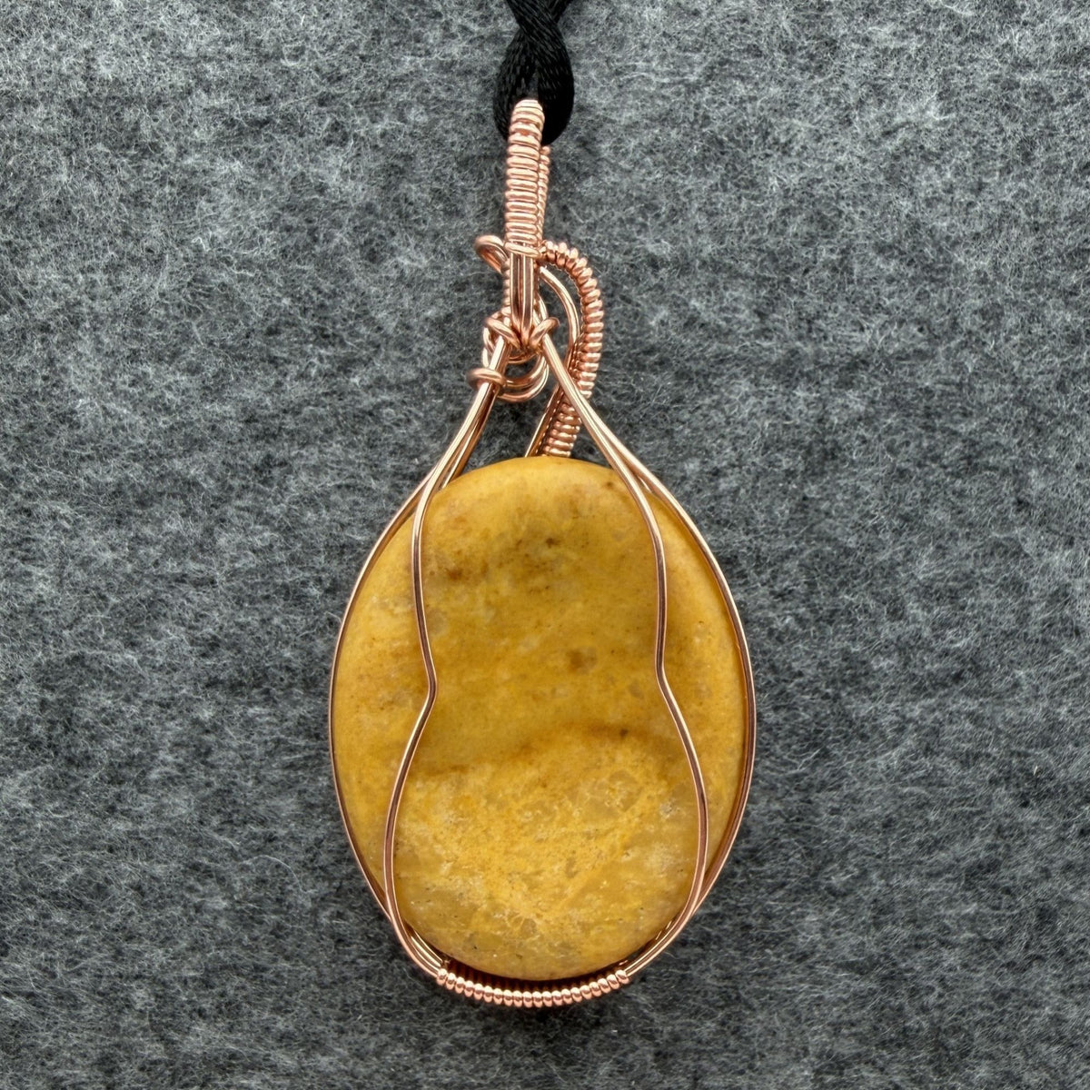 Back view of an oval-shaped yellow jasper stone pendant wrapped in copper wire with decorative swirls, hanging from a black cord against a textured gray background.