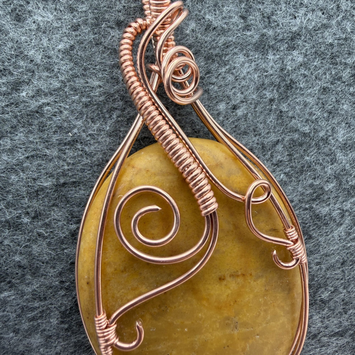 Detail view of an oval-shaped yellow jasper stone pendant wrapped in copper wire with decorative swirls, hanging from a black cord against a textured gray background.