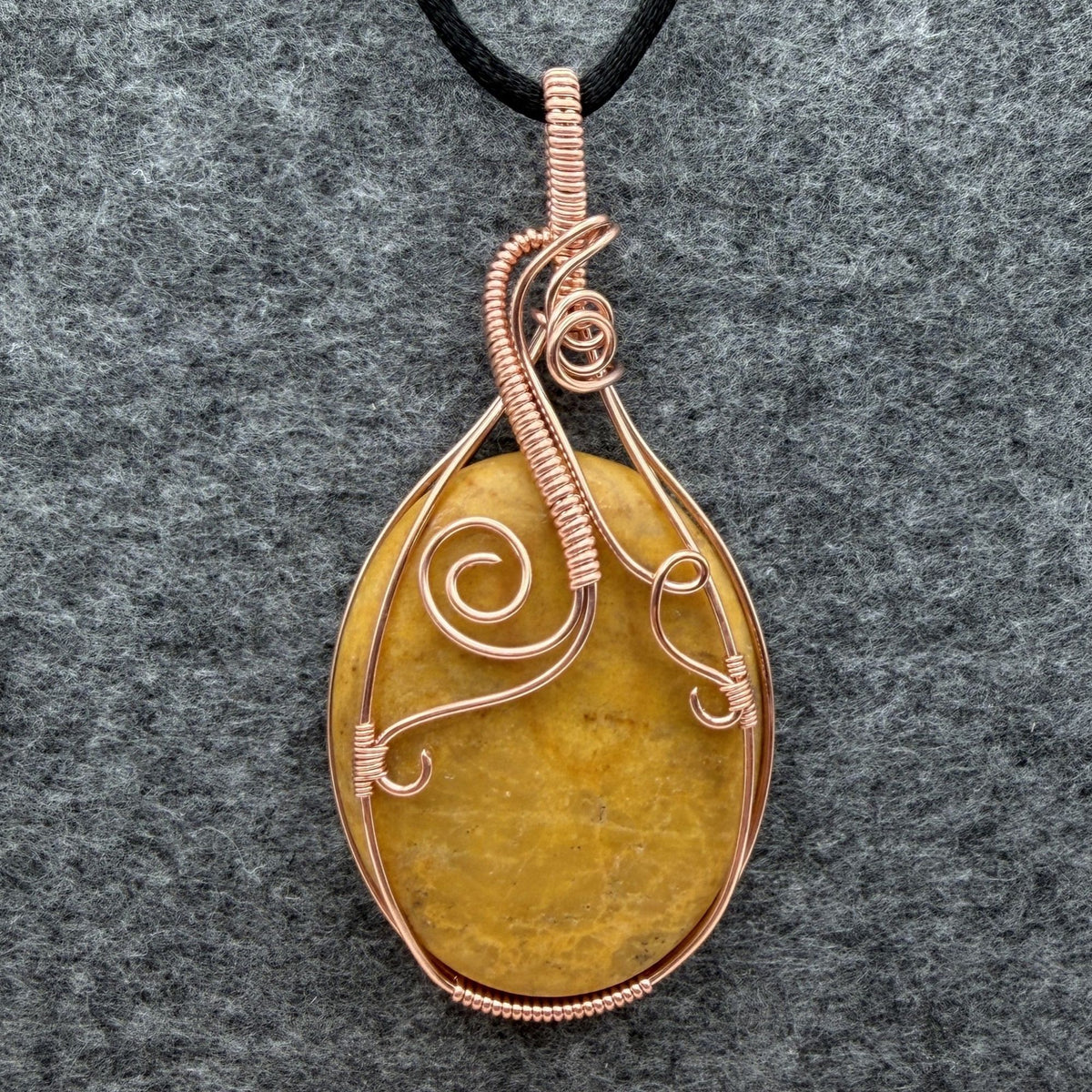 Front view of an oval-shaped yellow jasper stone pendant wrapped in copper wire with decorative swirls, hanging from a black cord against a textured gray background.