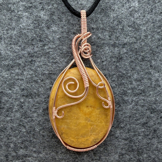 Front view of an oval-shaped yellow jasper stone pendant wrapped in copper wire with decorative swirls, hanging from a black cord against a textured gray background.