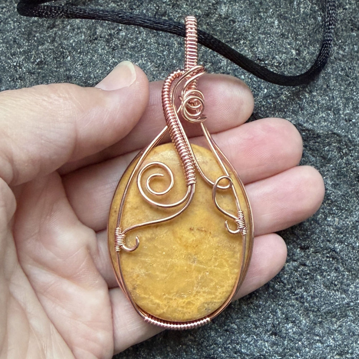 Front view of an oval-shaped yellow jasper stone pendant wrapped in copper wire with decorative swirls, hanging from a black cord held in the artist's hand.