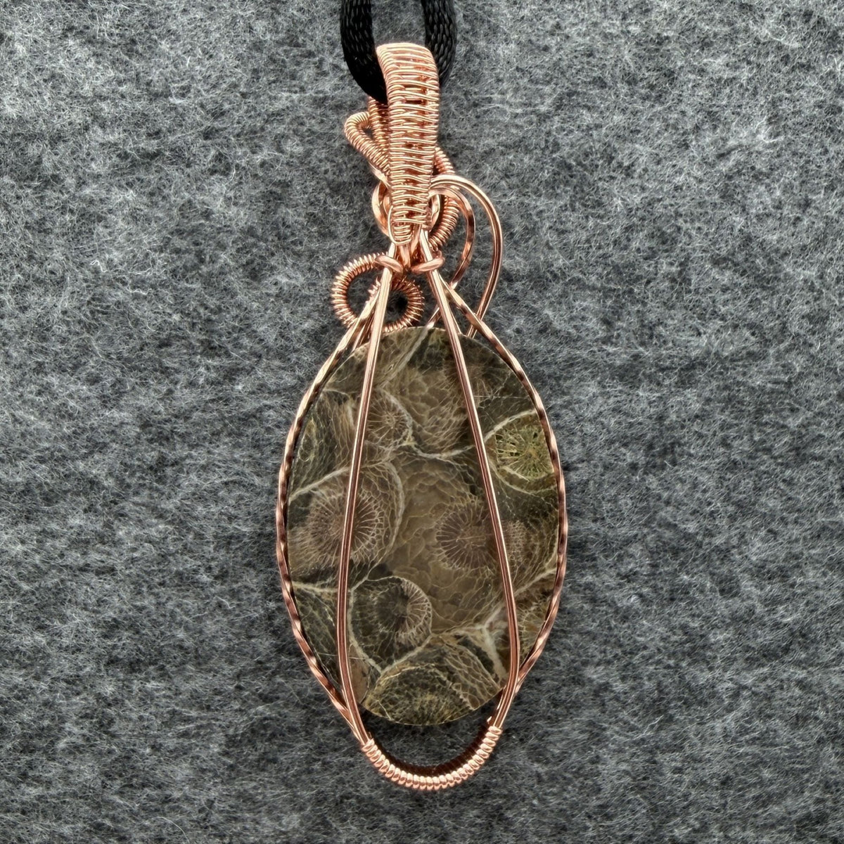 Back view of a handmade oval pendant necklace featuring a fossil coral cabochon wrapped in copper wire, suspended from a black cord against a gray felt background.