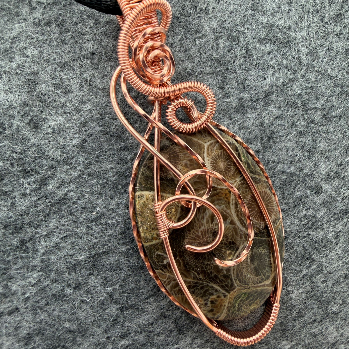 Detail view of a handmade oval pendant necklace featuring a fossil coral cabochon wrapped in copper wire, suspended from a black cord against a gray felt background.