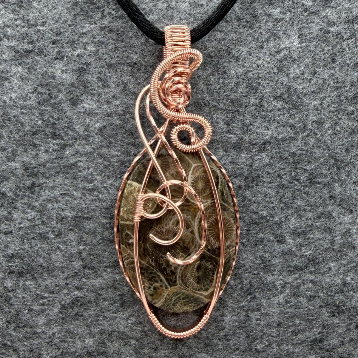 Front view of a handmade oval pendant necklace featuring a fossil coral cabochon wrapped in copper wire, suspended from a black cord against a gray felt background.
