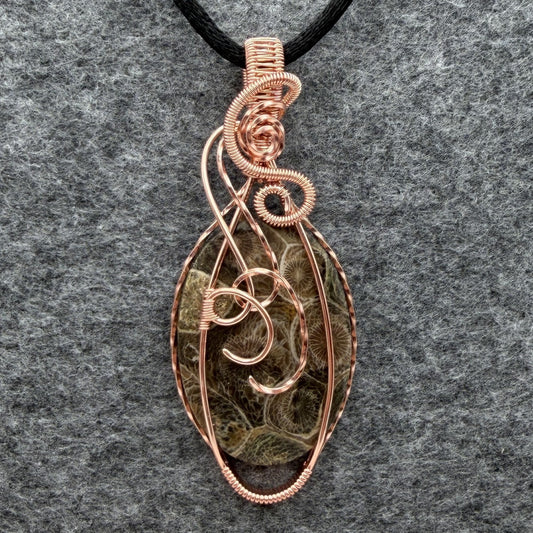 Front view of a handmade oval pendant necklace featuring a fossil coral cabochon wrapped in copper wire, suspended from a black cord against a gray felt background.