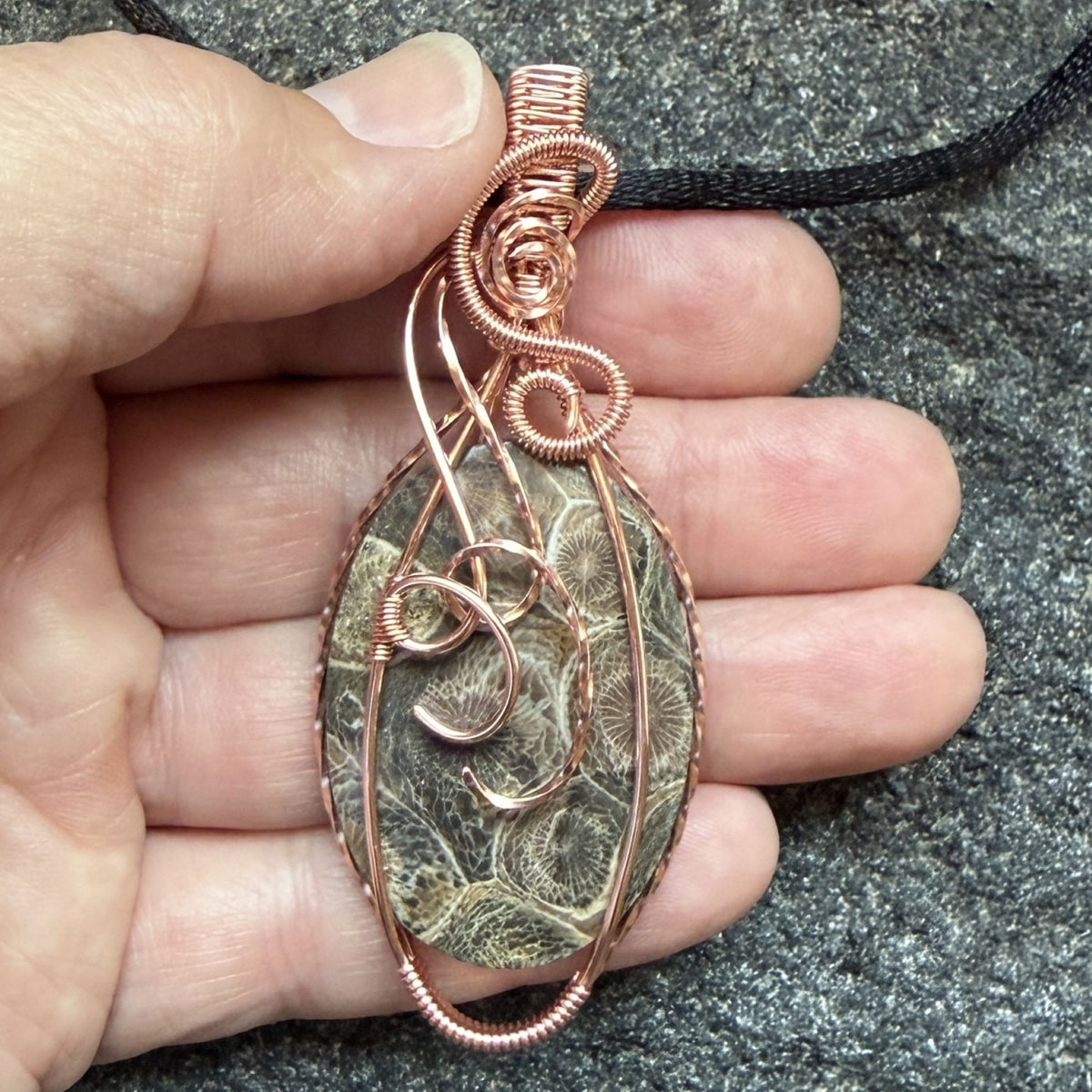 Front view of a handmade oval pendant necklace featuring a fossil coral cabochon wrapped in copper wire, suspended from a black cord held in the artist's hand.
