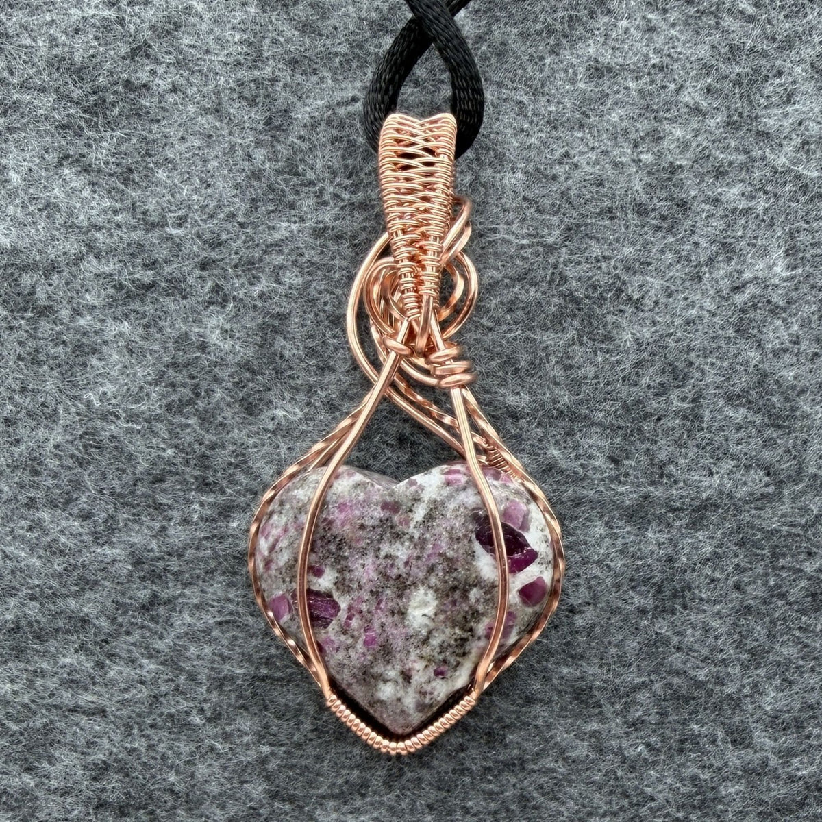 Back view of a heart-shaped stone pendant with pink ruby flecks, wrapped in copper wire with a black cord, displayed on a textured gray background.