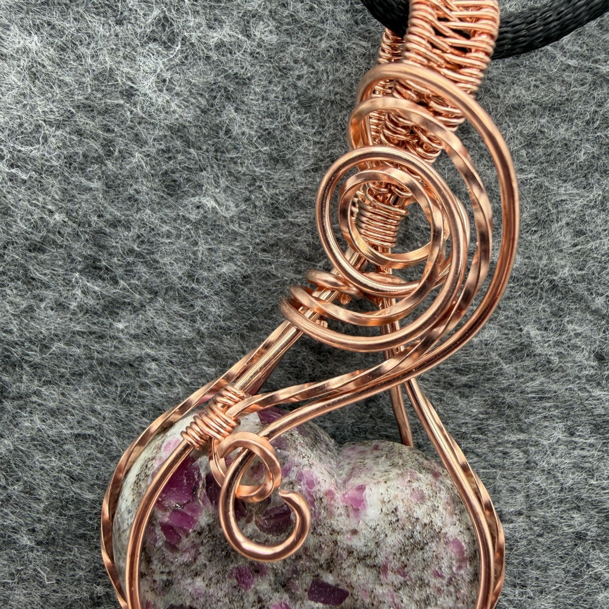 Detail view of a heart-shaped stone pendant with pink ruby flecks, wrapped in copper wire with a black cord, displayed on a textured gray background.
