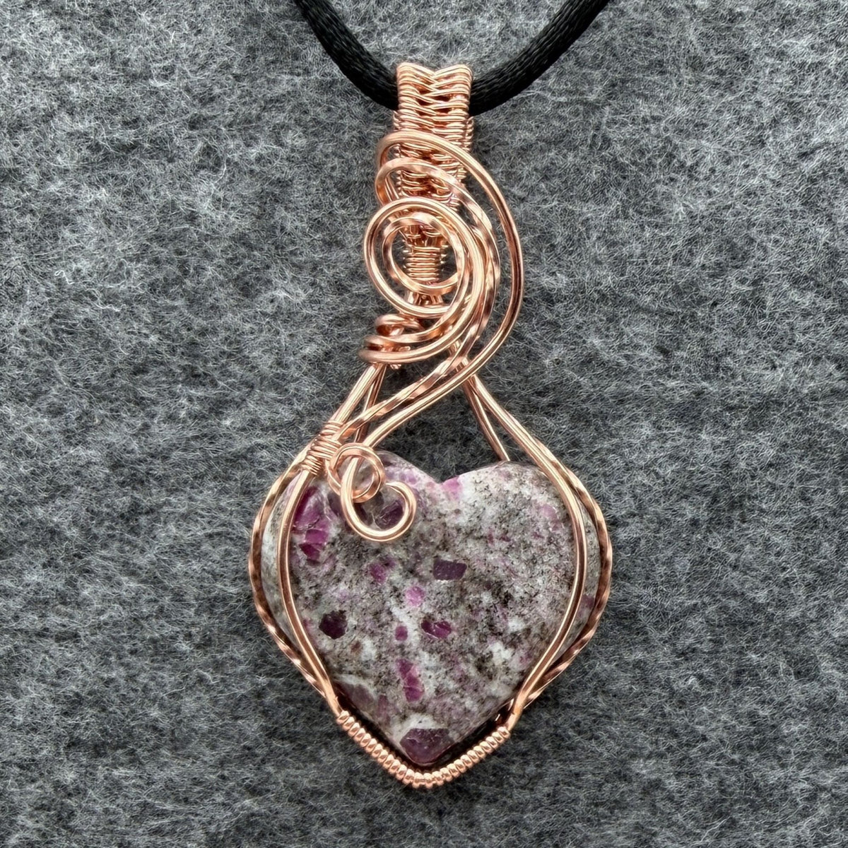 Front view of a heart-shaped stone pendant with pink ruby flecks, wrapped in copper wire with a black cord, displayed on a textured gray background.