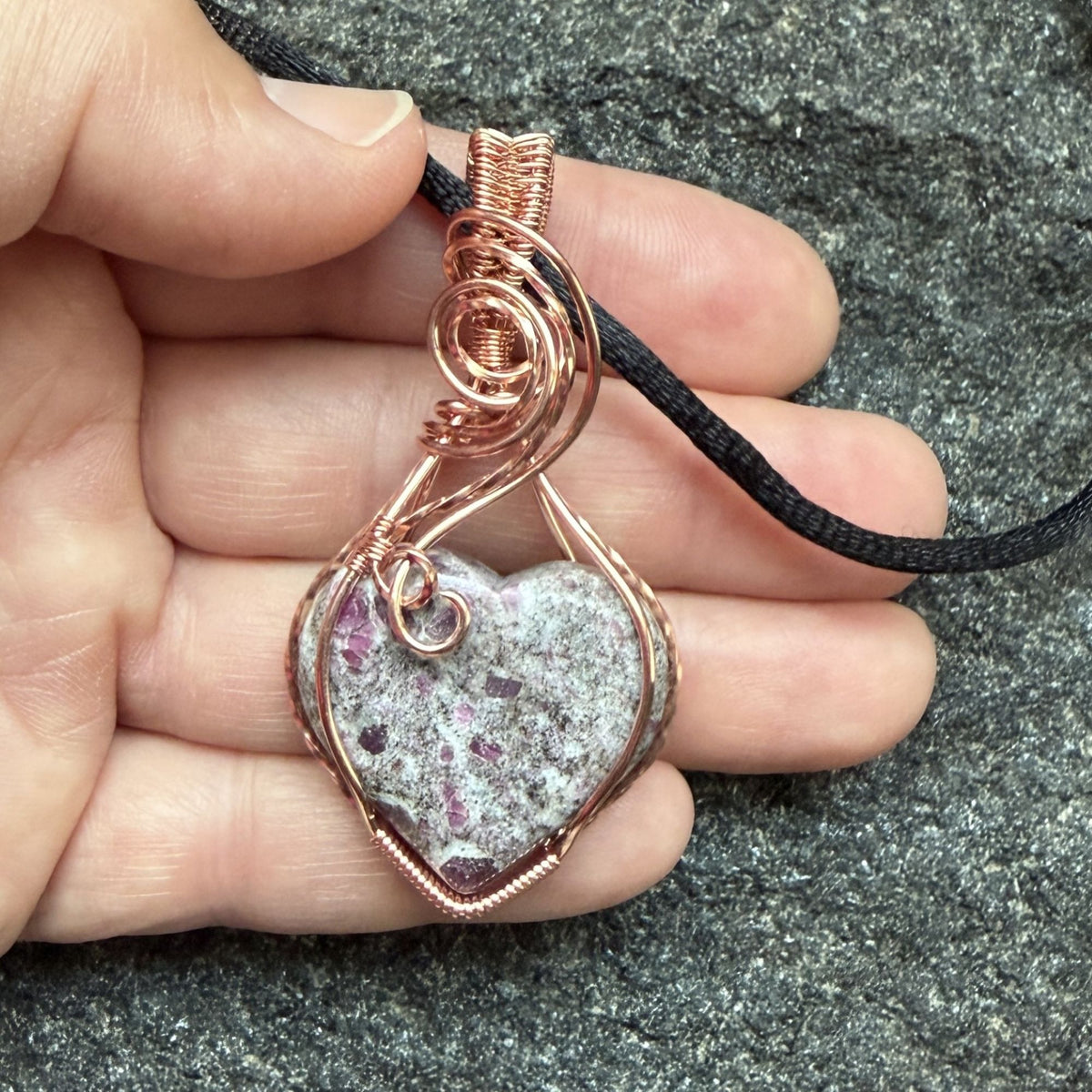 Front view of a heart-shaped stone pendant with pink ruby flecks, wrapped in copper wire with a black cord, displayed in the artist's hand.