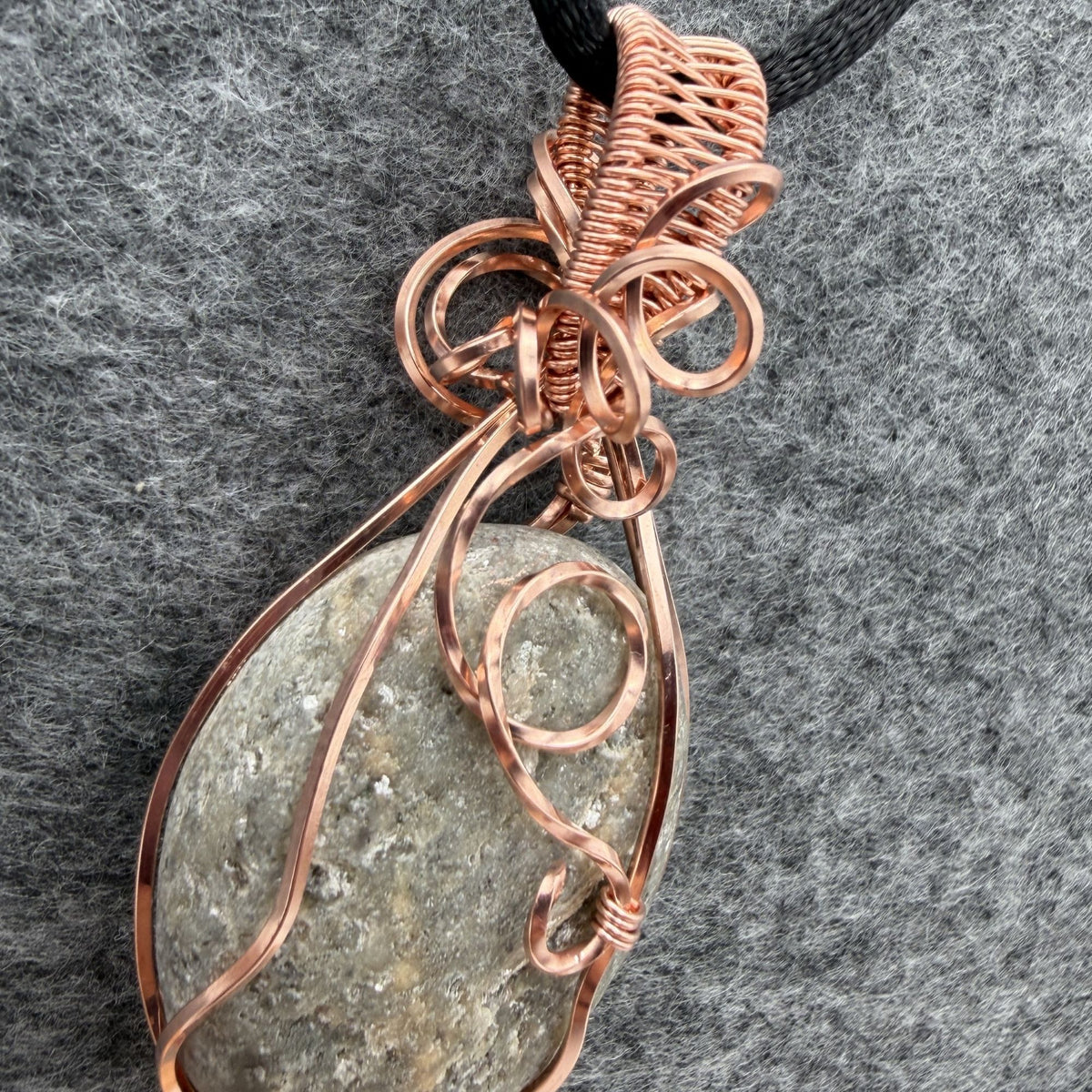 Detail view of side one of a reversible river stone pendant wrapped in copper wire and suspended on a black satin cord against a gray felt background.