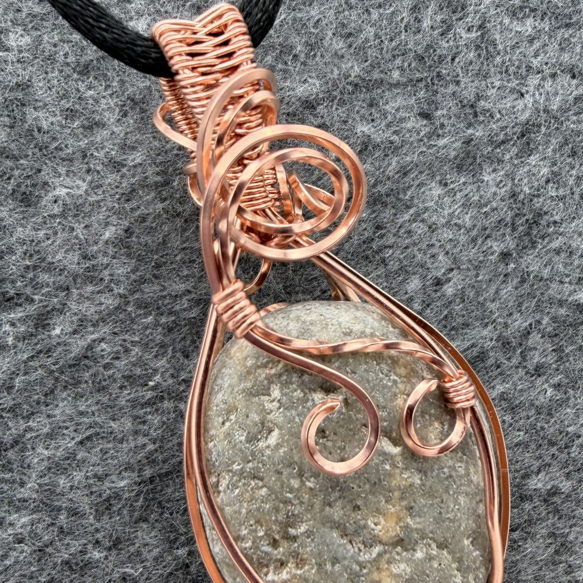 Detail view of side two of a reversible river stone pendant wrapped in copper wire and suspended on a black satin cord against a gray felt background.