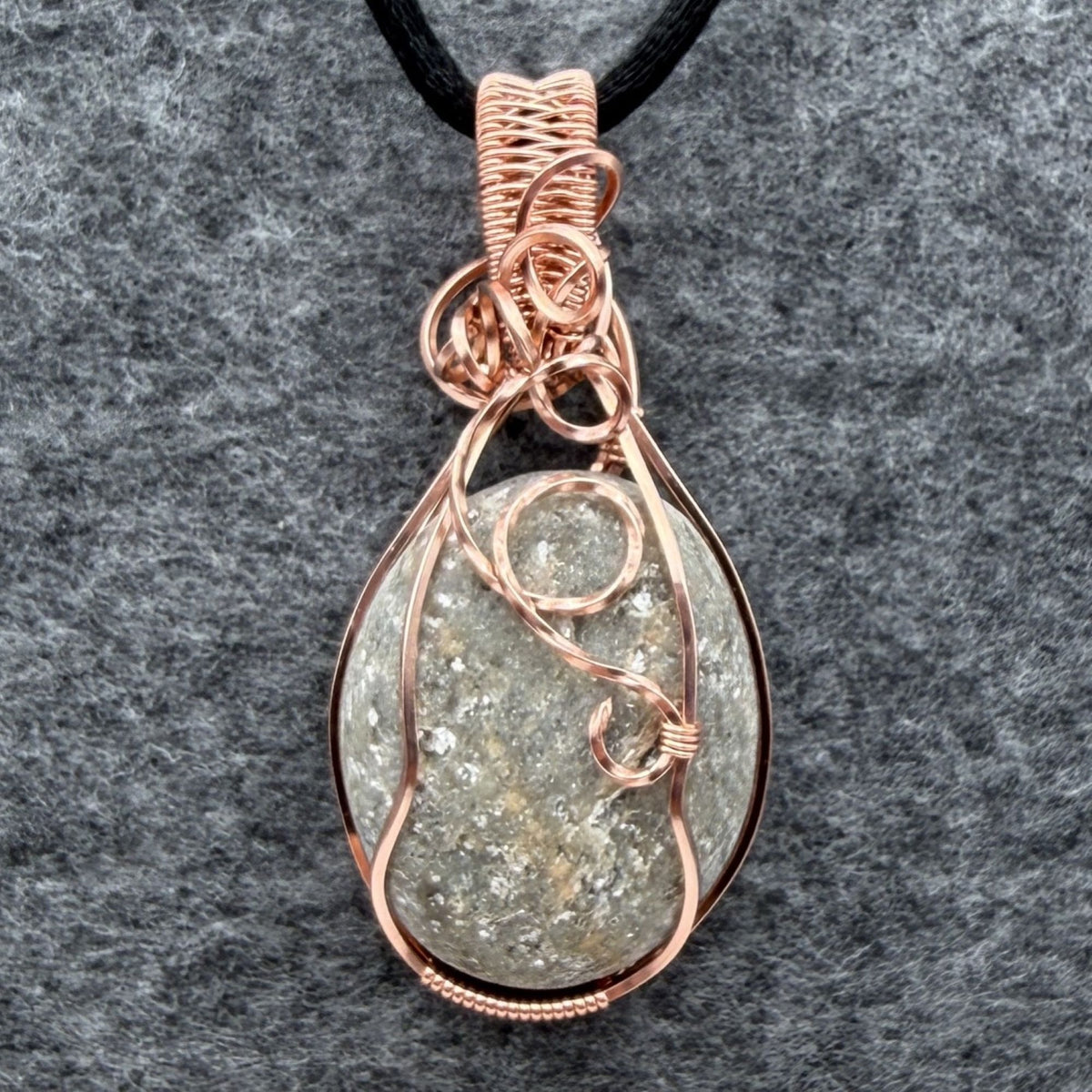 Front view of side one of a reversible river stone pendant wrapped in copper wire and suspended on a black satin cord against a gray felt background.