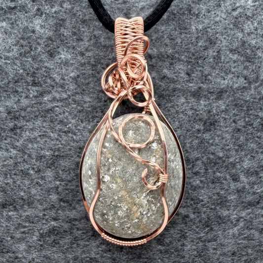 Front view of side one of a reversible river stone pendant wrapped in copper wire and suspended on a black satin cord against a gray felt background.