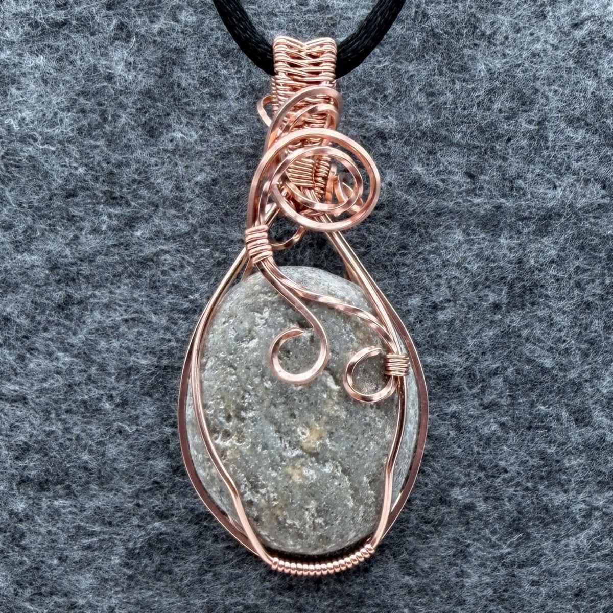 Front view of side two of a reversible river stone pendant wrapped in copper wire and suspended on a black satin cord against a gray felt background.