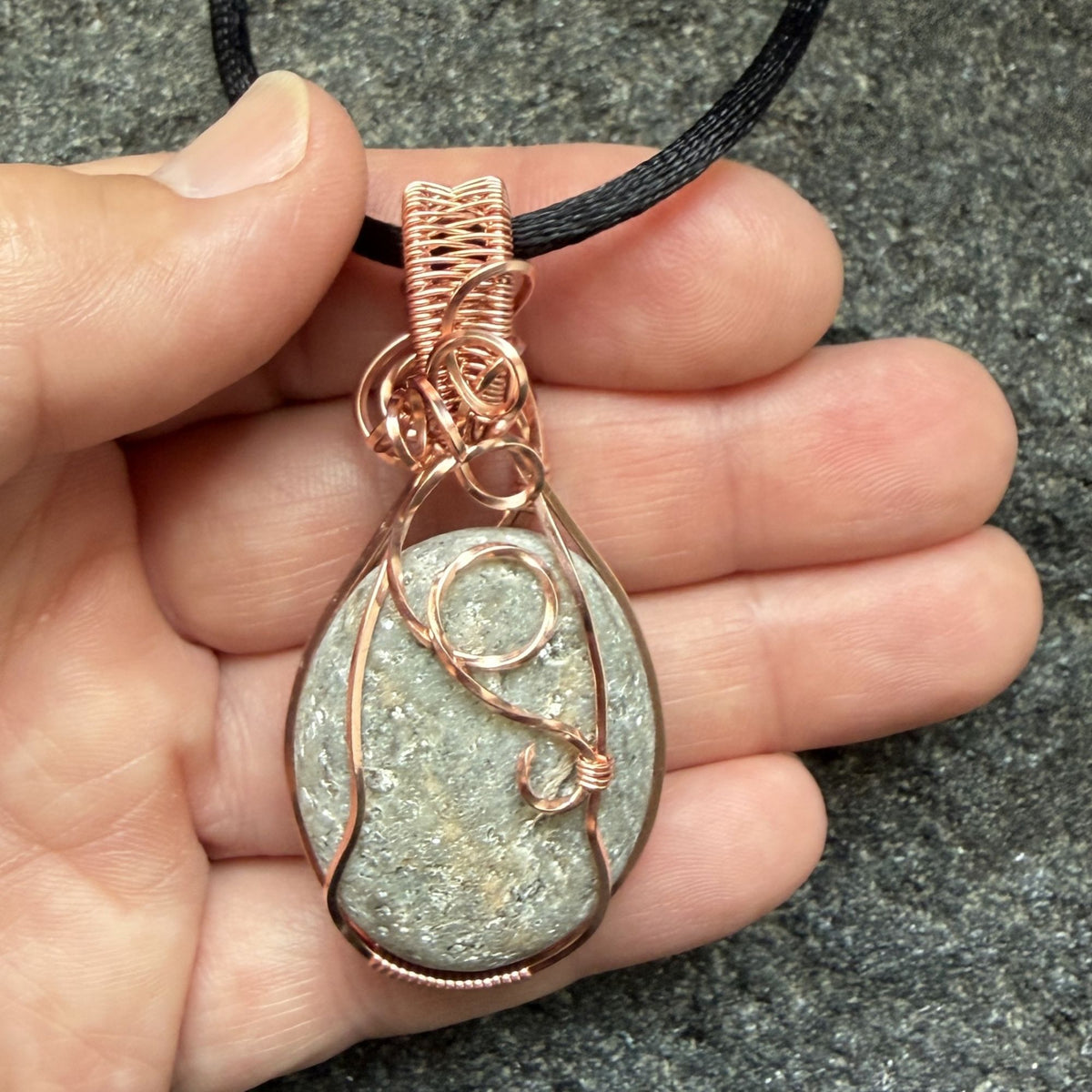 Front view of side one of a reversible river stone pendant wrapped in copper wire and suspended on a black satin cord held in the artist's hand.