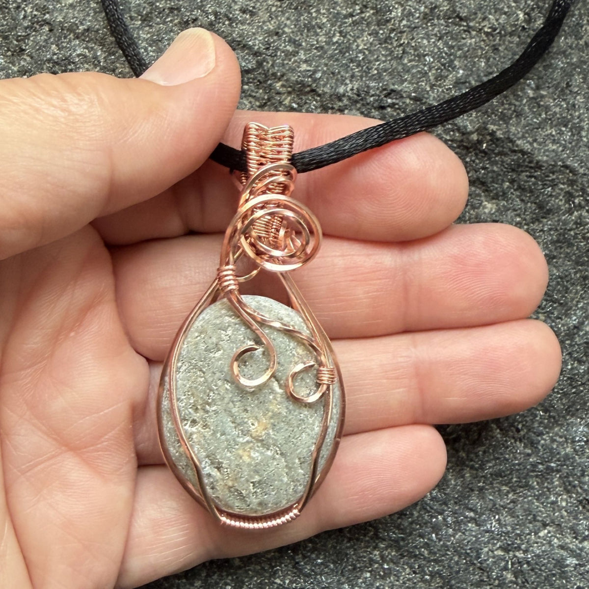 Front view of side one of a reversible river stone pendant wrapped in copper wire and suspended on a black satin cord held in the artist's hand.