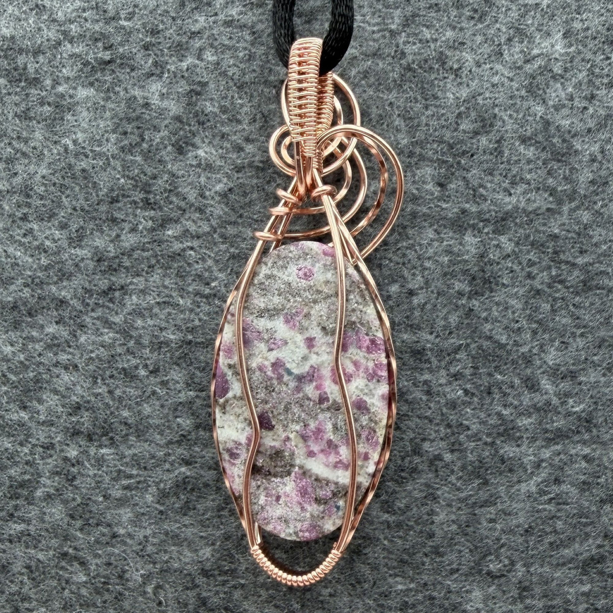 Back view of a handmade copper wire-wrapped pendant featuring a ruby albite stone with pink ruby in a gray and white speckled matrix, hanging from a black cord against a textured gray felt background.