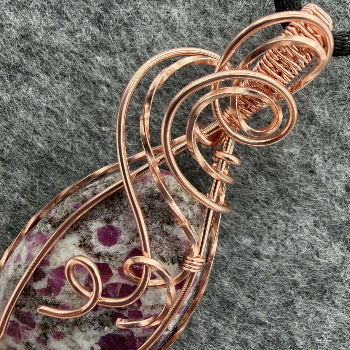 Detail view of a handmade copper wire-wrapped pendant featuring a ruby albite stone with pink ruby in a gray and white speckled matrix, hanging from a black cord against a textured gray felt background.