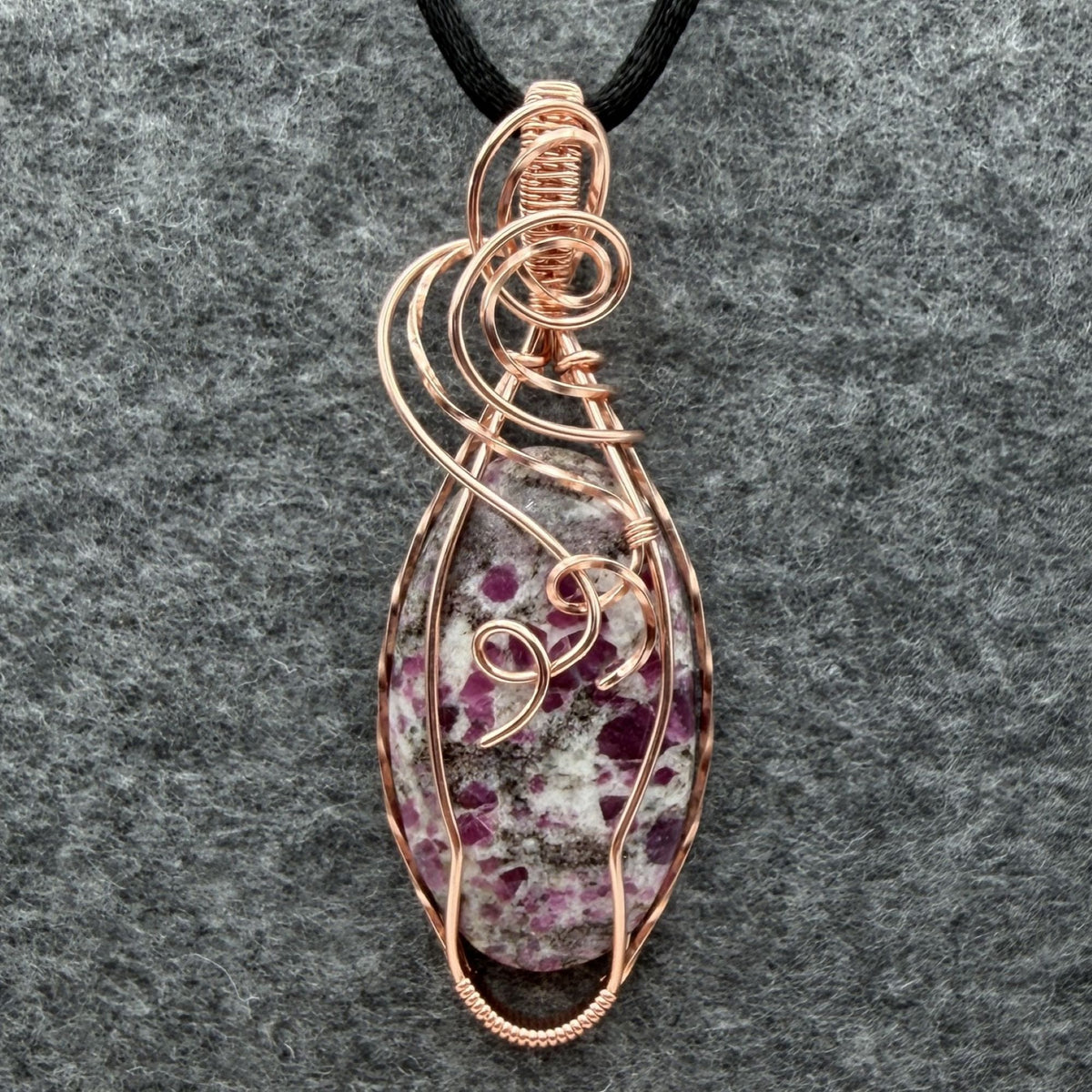 Front view of a handmade copper wire-wrapped pendant featuring a ruby albite stone with pink ruby in a gray and white speckled matrix, hanging from a black cord against a textured gray felt background.
