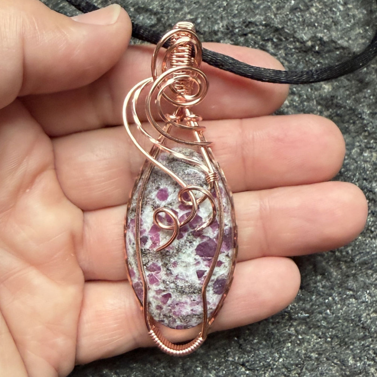 Front view of a handmade copper wire-wrapped pendant featuring a ruby albite stone with pink ruby in a gray and white speckled matrix, hanging from a black cord held in the artist's hand.