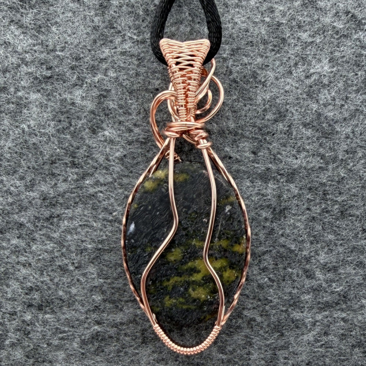 Back view of a teardrop-shaped wire-wrapped pendant necklace featuring a dark stone with olive green inclusions, set in copper wire and suspended from a black cord, shown against a textured gray background.