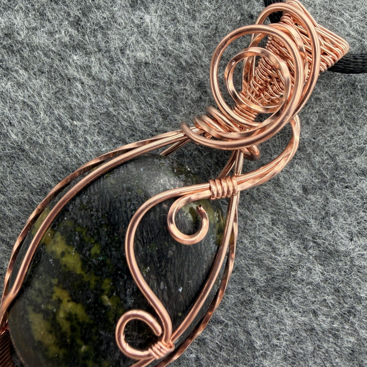 Detail view of a teardrop-shaped wire-wrapped pendant necklace featuring a dark stone with olive green inclusions, set in copper wire and suspended from a black cord, shown against a textured gray background.