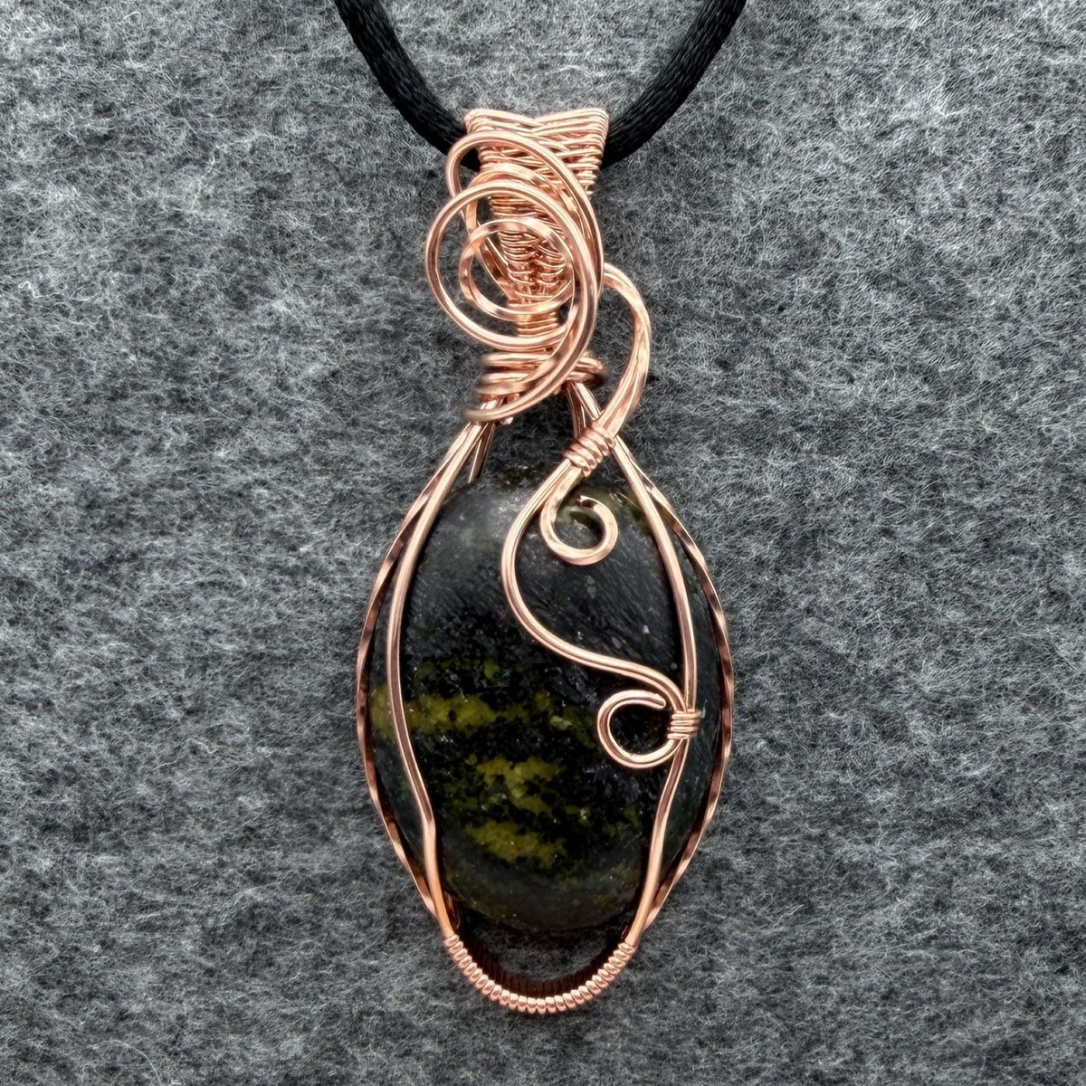 Front view of a teardrop-shaped wire-wrapped pendant necklace featuring a dark stone with olive green inclusions, set in copper wire and suspended from a black cord, shown against a textured gray background.
