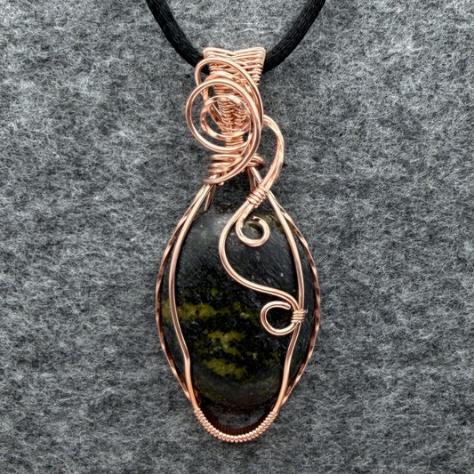 Front view of a teardrop-shaped wire-wrapped pendant necklace featuring a dark stone with olive green inclusions, set in copper wire and suspended from a black cord, shown against a textured gray background.