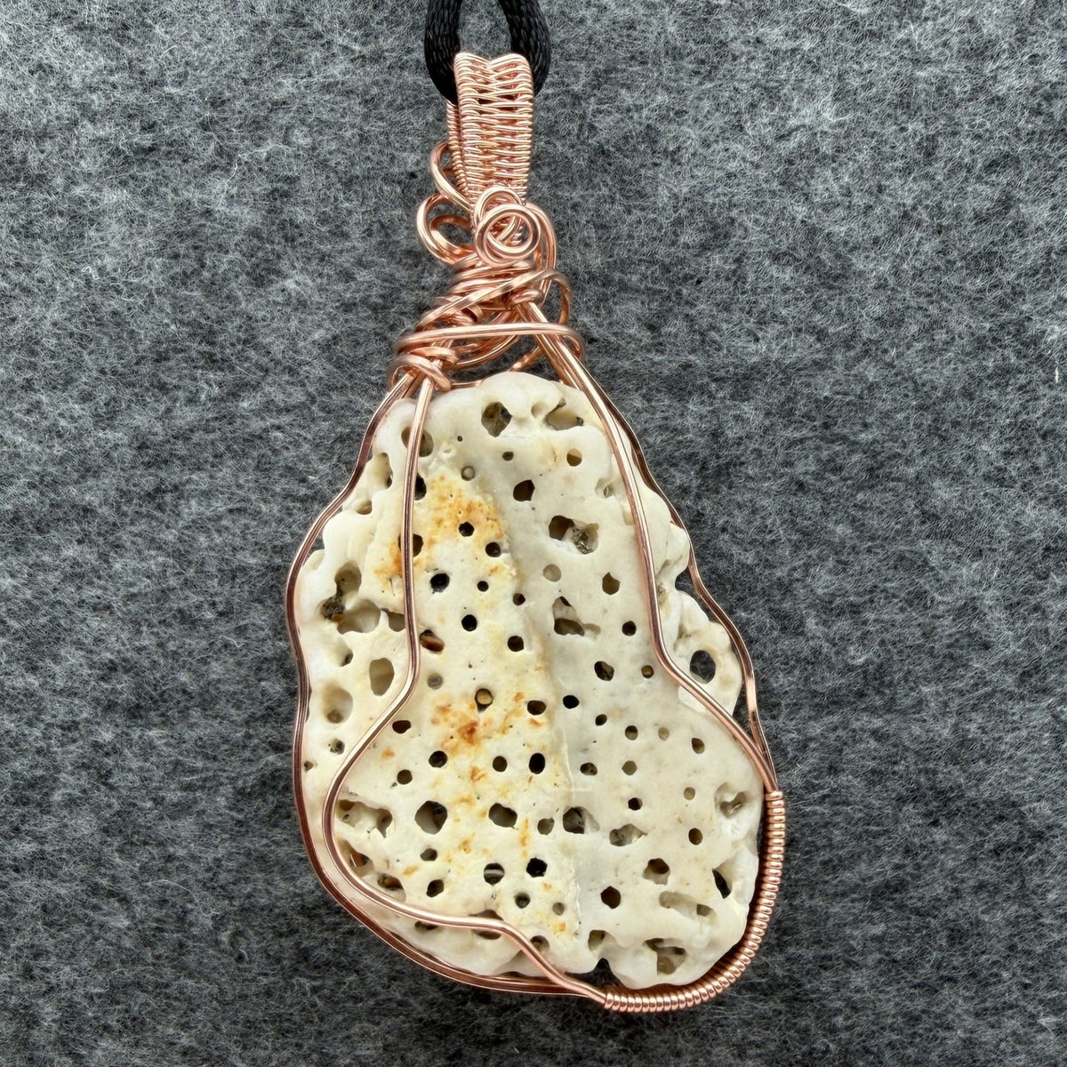 Back view of a teardrop-shaped porous cream shell pendant wrapped in copper wire, hanging from a black cord against a textured gray background.