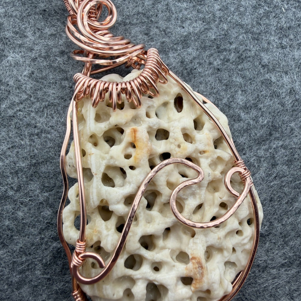 Detail view of a teardrop-shaped porous cream shell pendant wrapped in copper wire, hanging from a black cord against a textured gray background.