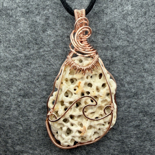 Front view of a teardrop-shaped porous cream shell pendant wrapped in copper wire, hanging from a black cord against a textured gray background.