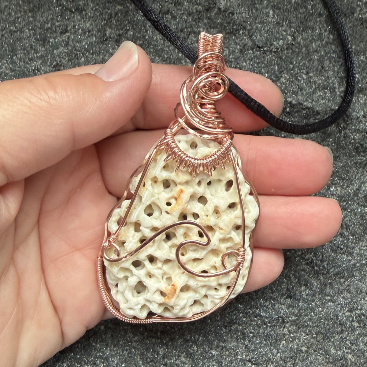 Front view of a teardrop-shaped porous cream shell pendant wrapped in copper wire, hanging from a black cord held in the artist's hand.