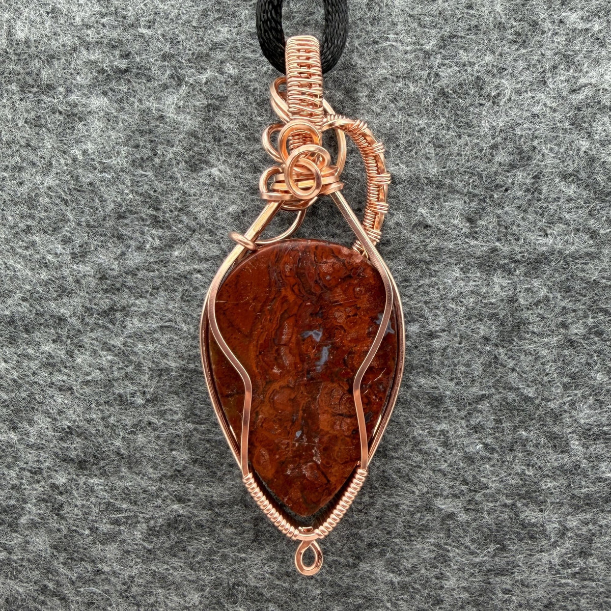 Back view of an inverted-teardrop shaped red plume agate pendant wrapped in copper wire and suspended from a black cord, shown against a textured gray background.