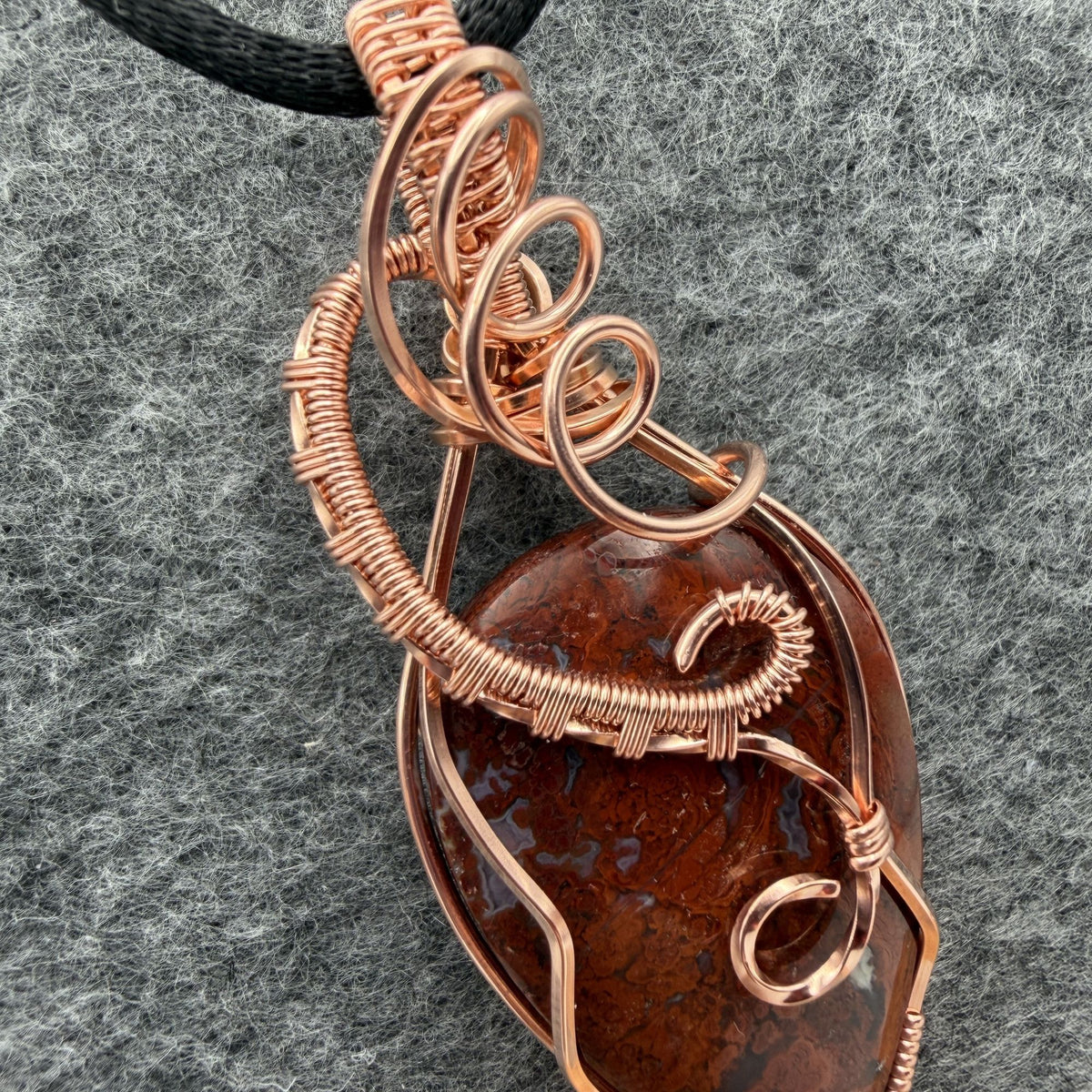 Detail view of an inverted-teardrop shaped red plume agate pendant wrapped in copper wire and suspended from a black cord, shown against a textured gray background.