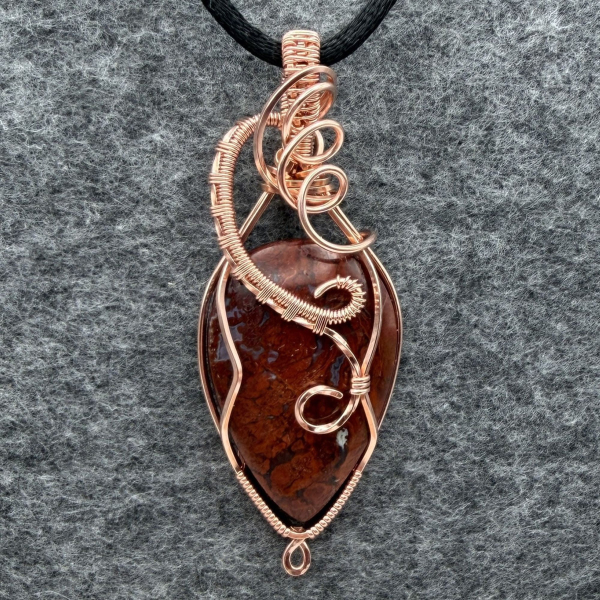 Front view of an inverted-teardrop shaped red plume agate pendant wrapped in copper wire and suspended from a black cord, shown against a textured gray background.